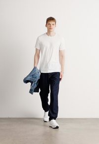 Hvit t-skjorte, marineblå bukser og hvite sneakers. Modellen holder en blå mønstrete skjorte. Enkelt design, avslappet passform og uformell stil.