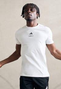 Bílá sportovní tričko s krátkými rukávy, s černým logem Adidas a textem "LES MILLS" na přední straně. Hladká tkanina, přiléhavý střih.