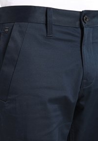 Tmavě modré kalhoty z hladké látky s knoflíkovým zapínáním, poutky na pásek a úzkým střihem s minimálními viditelnými švy.