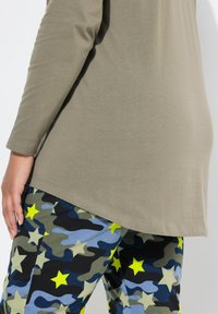 Olivi vihreä pitkähihaisen paita, jossa on kaareva helma, yhdistettynä tummiin maastohousuihin, joissa on sinisiä, vihreitä ja kirkkaan keltaisia tähtikuvioita.