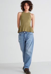 Top peplum verde oliva con acabado texturizado, combinado con jeans desgastados azul claro. Modelo lleva zapatos marrones. Fondo blanco.
