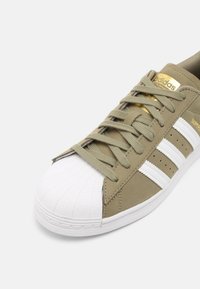 Beige Adidas Superstar-sneakers med vit snäppa, vita ränder, beige snören och guld Adidas-logotyp på tungan och sidan.