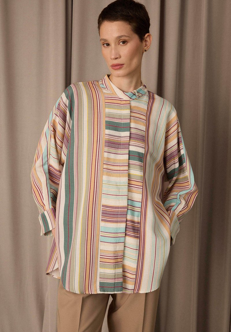 Personne portant une chemise ample à manches longues rayée avec des lignes verticales multicolores et un pantalon beige, debout devant un fond de rideau beige uni.
