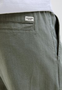 Pantalones de tela verdes con un bolsillo trasero de ojal y una etiqueta blanca de la marca Jack & Jones cosida encima del bolsillo.