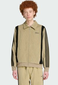Beige corduroy jacket with black side stripes and zip closure. Features a collared neck and embroidered branding on the chest.