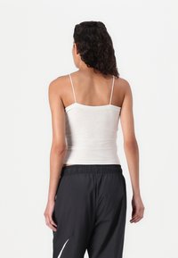 Camisola branca de algodão com alças finas, silhueta ajustada e textura suave, combinada com calças de treino pretas com um logo branco.