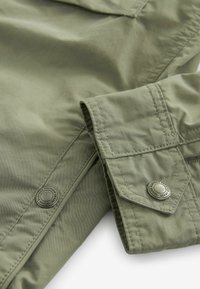 Tissu de veste léger en vert olive avec une texture lisse. Les manchettes sont dotées d'une fermeture à bouton et de coutures décoratives. Le profil comprend un rabat de poche.