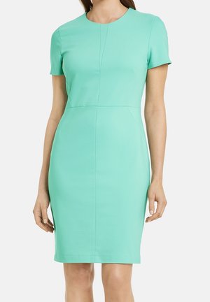 Femme portant une robe ajustée, longueur genou, manches courtes, turquoise clair uni avec un col rond et des détails subtils de coutures.