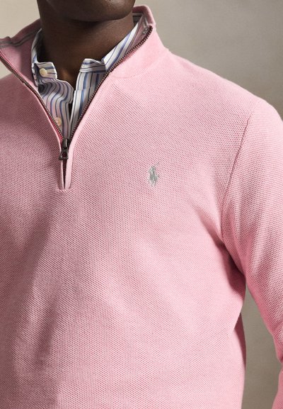 Ανοιχτό ροζ πουλόβερ με υφή πλεκτού, με γιακά μισής ζώνης και διακριτικό λογότυπο στο στήθος. Κάτω από αυτό, διακρίνεται ένα ριγέ πουκάμισο με γιακά.