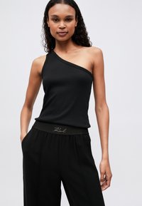 Fekete, bordázott, egy vállon pántos trikó. Laza fazonú fekete nadrággal párosítva, amelynek elasztikus derékrésze és diszkrét logó részletei vannak.