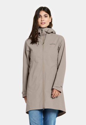 Woman wearing a beige Didriksons raincoat with hood, standing with hands partly in pockets, set against a plain light background.