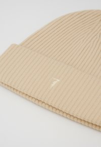 Gorro de punto beige acanalado con puño doblado y un logo de rayo bordado en un tono más claro en la parte frontal.