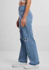 Svetlomodré rifle s širokými nohavicami, s rozstrapkanými okrajmi a viacerými poškodenými miestami. Kombinované s bielymi teniskami, zobrazené v bočnom pohľade.