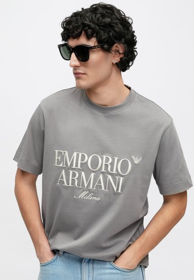 T-shirt cinzenta de algodão com mangas curtas, com a inscrição "EMPORIO ARMANI Milano" em relevo de cor creme, complementada por um pequeno emblema com o logótipo.