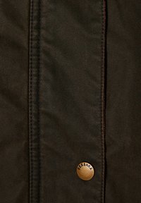 Mørkebrunt stof med en glat tekstur, der har en fremtrædende messingknap mærket "BERSHKA" og synlig syning langs sømmen.