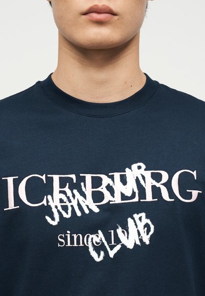 Κοντινό πλάνο ενός ατόμου που φοράει μπλε σκούρο πουκάμισο με ανάμεικτο λευκό και ροζ κείμενο που γράφει "ICEBERG JUNIOR CLUB since 1974".