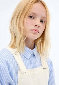 Lichtblauw gestreept overhemd met kraag onder beige tuinbroek met knoopdetails. Model heeft golvend blond haar en natuurlijke make-up, waardoor sproeten zichtbaar zijn.