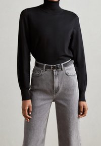 Schwarzer Rollkragenpullover kombiniert mit hoch geschnittenen grauen Jeans und einem grauen Gürtel. Die Jeans haben ein klassisches Fünf-Taschen-Design und gerades Bein.