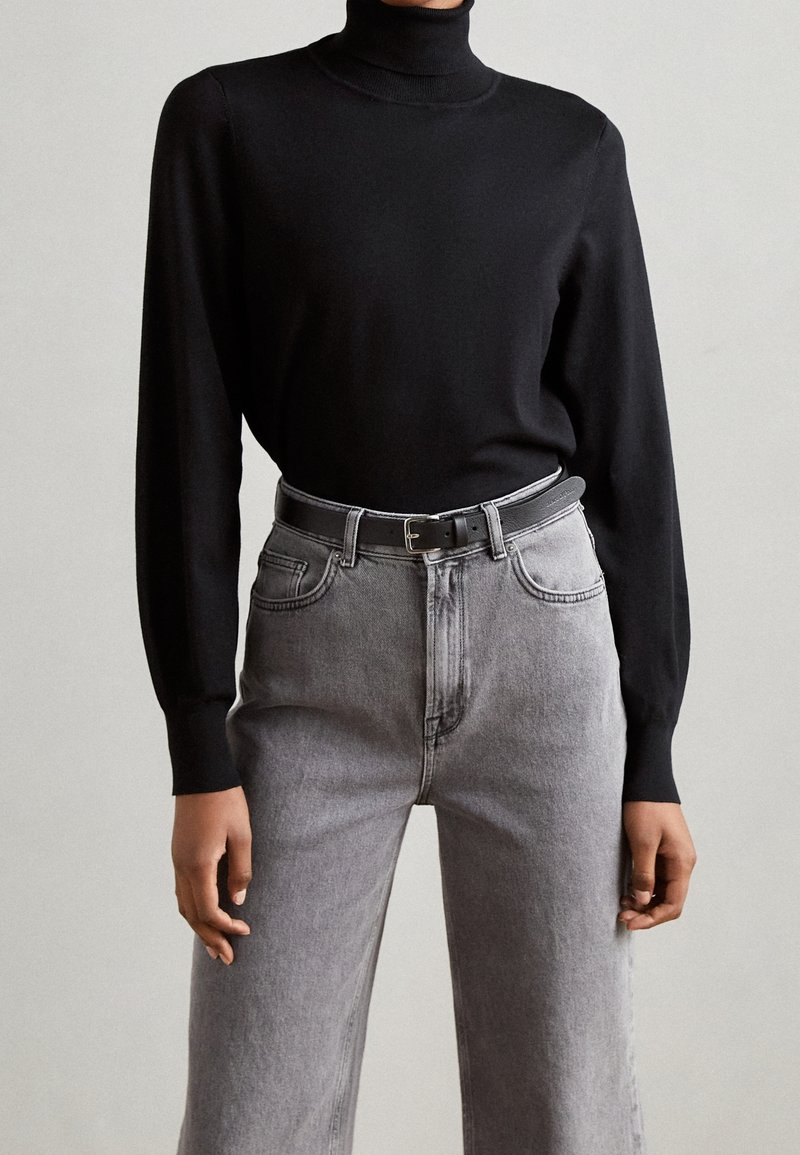 Schwarzer Rollkragenpullover kombiniert mit hoch geschnittenen grauen Jeans und einem grauen Gürtel. Die Jeans haben ein klassisches Fünf-Taschen-Design und gerades Bein.
