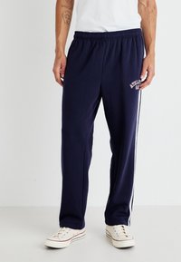 Marineblå joggebukser laget av myk stoff, med elastisk midje, hvite sidelister og et trykt logo nær lommen.