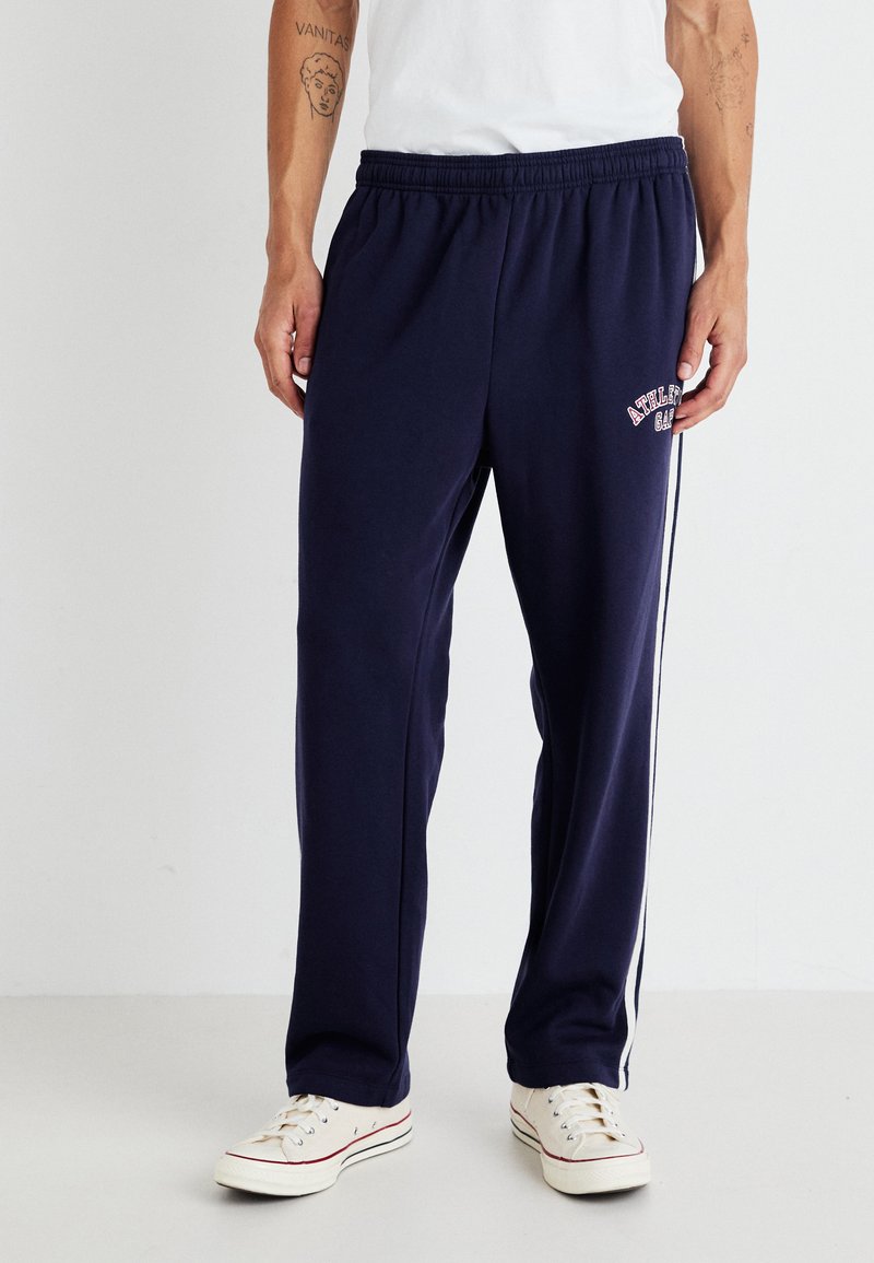 Marineblå joggebukser laget av myk stoff, med elastisk midje, hvite sidelister og et trykt logo nær lommen.