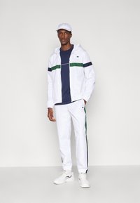 Conjunto atlético masculino com uma jaqueta branca com fecho zip e riscas verdes e azuis, combinada com calças brancas e um boné branco.