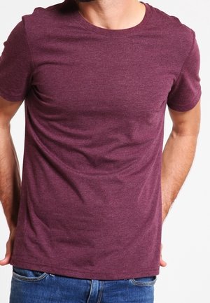 Mann trägt eng anliegendes kurzärmeliges bordeauxrotes T-Shirt und blaue Jeans vor weißem Hintergrund, Oberkörper sichtbar, Hände hinter dem Rücken.