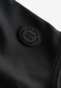 Musta kangas, jossa on sileä pinta ja pyöreä, kohokuvioitu musta logolaatta, jossa lukee "STAY ACTIVE" ja geometrisia koristeita.