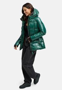 Teal puffer jack met een glanzende afwerking, gequilt ontwerp en een capuchon. Gecombineerd met zwarte jeans en zwarte sneakers.