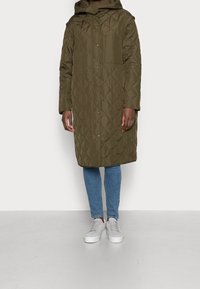 Cappotto lungo imbottito verde oliva con cappuccio, dotato di chiusura a scatto frontale e tasca sul petto. Indossato sopra jeans azzurri e sneakers bianche.