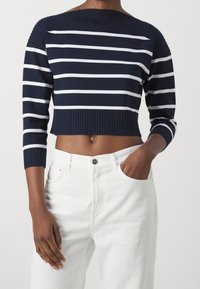 Maglione cropped a righe blu navy e bianche con design off-shoulder, scollo a costine e maniche a tre quarti, abbinato a jeans bianchi a vita alta.