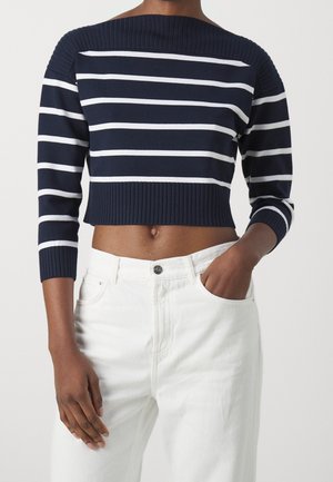 Navy and white striped cropped sweater with off-shoulder design, ribbed neckline, and three-quarter sleeves, paired with white high-waisted jeans.