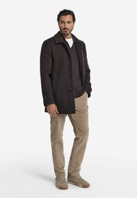 Mörk ullrock med spetsig krage, knappstängning och påsydda fickor, i kombination med en mörk tröja, beige manchesterbyxor och beige sneakers.