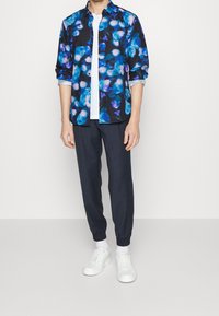 Camisa multicolorida con patrones florales azules y morados sobre un fondo negro, combinada con pantalones oscuros y zapatillas blancas.