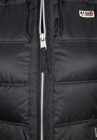 Schwarze Pufferjacke mit grauem Reißverschluss, mit gesteppten Abschnitten und einem kleinen Emblem auf der oberen linken Seite. Reißverschluss vorne.