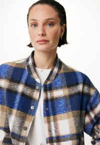 Modrý a pískový kostkovaný kabát s kulatým výstřihem, zapínáním na patentky a měkkou texturou, vrstvený přes bílou košili.