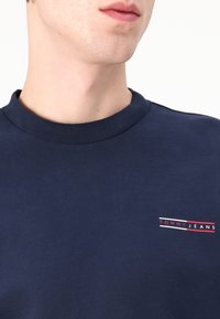 Tamno plavi dukserica s rebrastim okruglim izrezom, s malim crvenim i bijelim "TOMMY JEANS" logotipom izvezenim na prsima. Mekana tekstura.