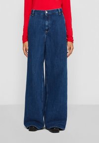 Person med høy midje, vid ben stil med mørkeblå denimjeans og en langermet rød topp, som står mot en lys, enkel bakgrunn.
