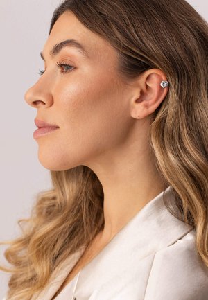 Frau mit langen, welligen Haaren in einer weißen Bluse, die einen silbernen, blumenförmigen Ohrcuff am oberen rechten Ohr trägt und im Profil nach links schaut.