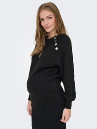 Nėščia moteris dėvi juodą megztinį su sidabriniais mygtukais ant peties ir juodą klostuotą sijoną, stovinti priešais vientisą foną.