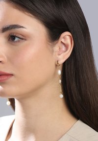 Goldene Creolenohrringe verfügen über hängende Stränge mit weißen Perlen und goldenen Akzenten. Das Design ist länglich und schafft ein zartes Erscheinungsbild.