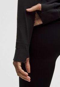 Fekete sportos, hosszú ujjú felső, hüvelykujjtartókkal, sima textúrával és testhezálló fazonnal, fekete leggingszel párosítva.