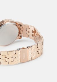 Närbild av ett klockarmband i roséguld med länkad armband och spänne som är graverat med "GUESS" på en vit bakgrund.