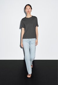T-shirt grigia a maniche corte con scollatura rotonda, abbinata a jeans dritti blu chiaro e sandali piatti marroni, su uno sfondo semplice.