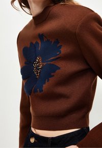 Pull court marron avec une grande fleur texturée bleu marine et des embellissements en perles, porté avec un jean bleu foncé.