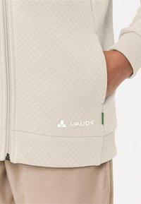 Veste zippée matelassée grise avec un logo blanc, poches latérales et poignets côtelés. Le tissu est doux avec un design texturé.