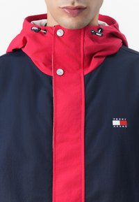 Marineblå og rød vandtæt jakke med høj krave, sølvfarvede knaplukninger og et lille "Tommy Jeans" logo på brystet.