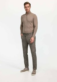 Hellbrauner Reißverschluss-Pullover mit langen Ärmeln, kombiniert mit gemusterten, figurbetonten dunkelgrauen Hosen und grauen Schnürsneakern.