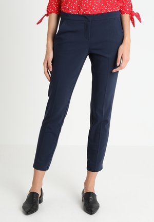 Mujer con pantalones azul marino ajustados hasta el tobillo, blusa roja con lunares blancos y zapatos negros sin cordones de punta afilada, posando frente a un fondo blanco.