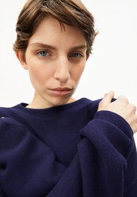 Marineblauwe gebreide trui met een brede bootvormige halslijn en een zachte textuur. De trui heeft omgeslagen mouwen en een close-up weergave van de halslijn.